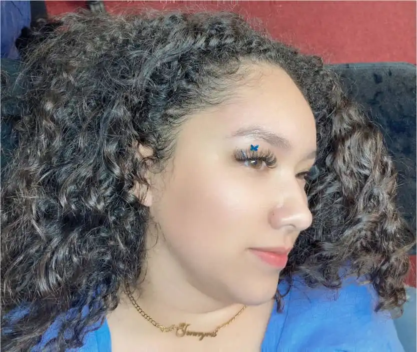 Woman with curly hair and eyelash extensions, wearing blue top and gold necklace.