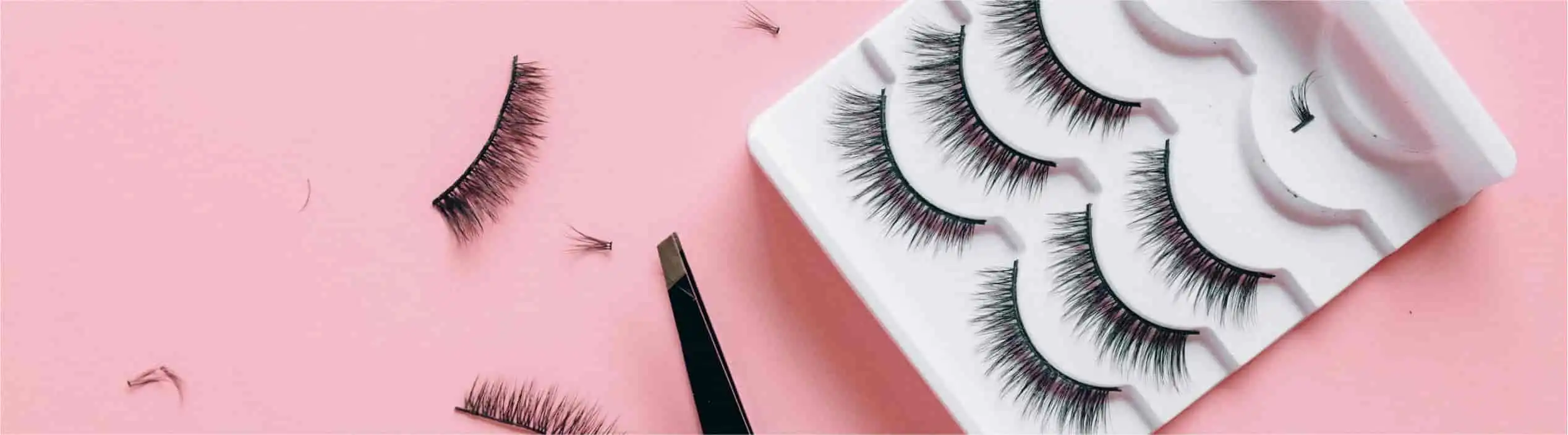 False eyelashes and applicator on pink background. White tray with multiple lash pairs, individual lashes beside it.